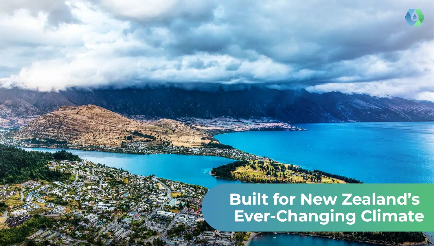 Aerial view of a New Zealand town beside a blue lake with mountains and dark clouds that show NZ climate variety